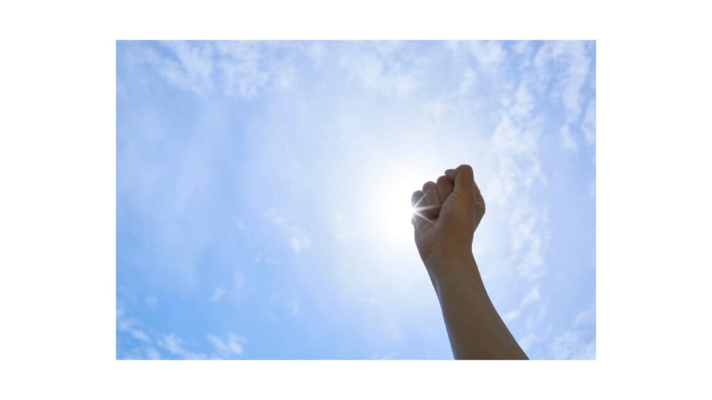 青空に向かって力強く拳を突き上げる腕のシルエット。太陽の光を浴びながら、前向きなエネルギーを感じさせる構図。