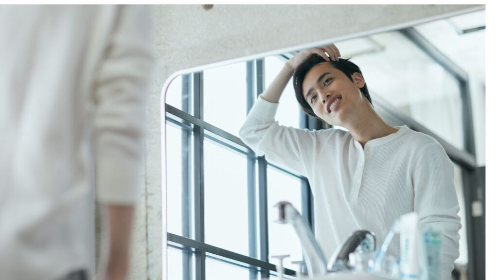 笑顔で洗面所の鏡を見つめる若い男性。清潔感のある白い服を着て、明るい空間でリラックスした様子。
