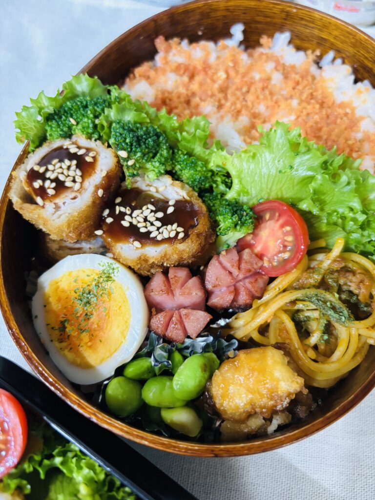 「曲げわっぱと黒色弁当箱に詰めた、鮭ふりかけご飯・ロールカツ・ゆで卵・枝豆・ミートスパ・ウインナー・ブロッコリー・レタス・ミニトマト・イカの天ぷらのお弁当。彩りがよく、木のわっぱが食材を引き立てている。」
