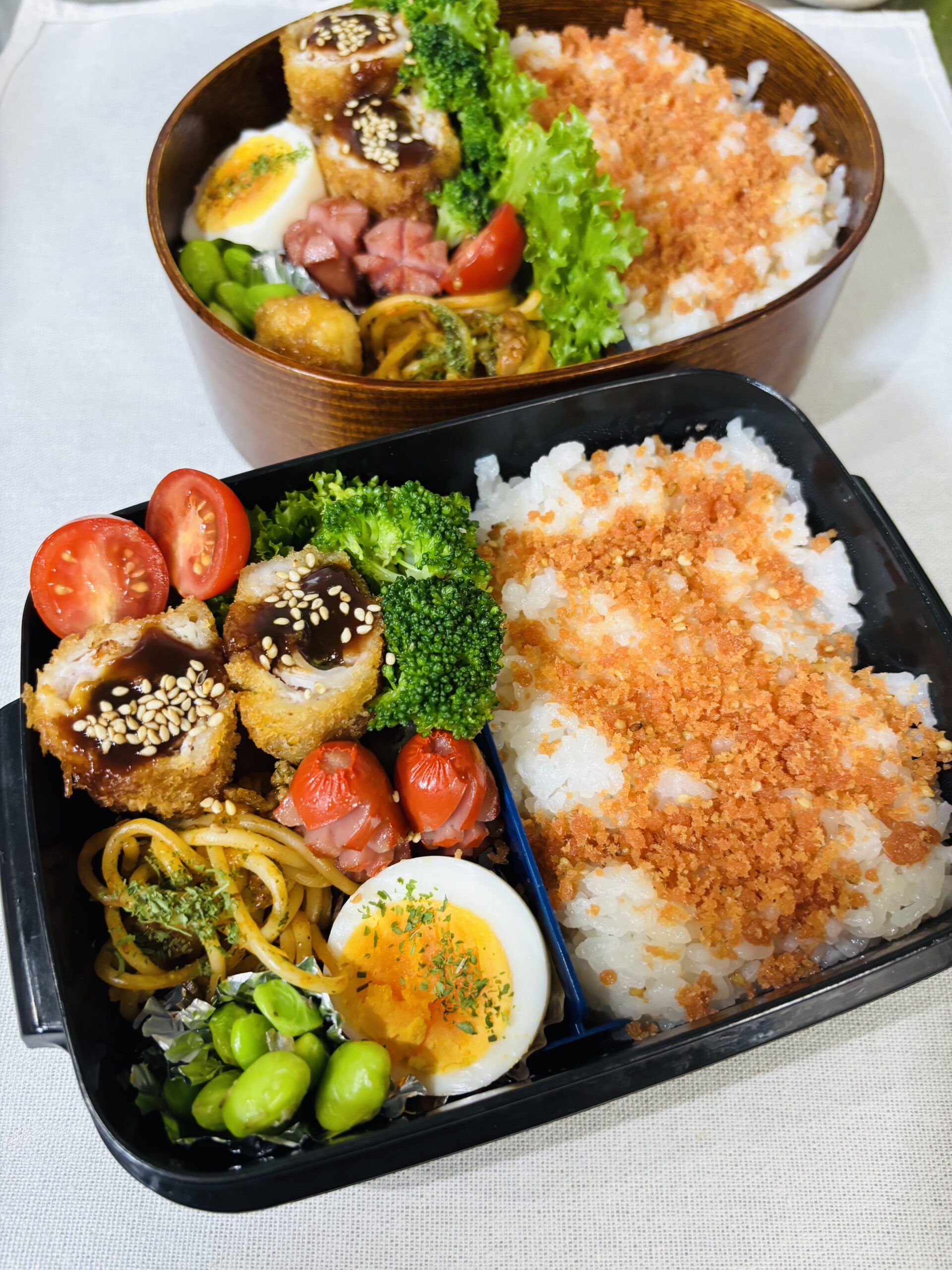 「曲げわっぱと黒色弁当箱に詰めた、鮭ふりかけご飯・ロールカツ・ゆで卵・枝豆・ミートスパ・ウインナー・ブロッコリー・レタス・ミニトマト・イカの天ぷらのお弁当。彩りがよく、木のわっぱが食材を引き立てている。」